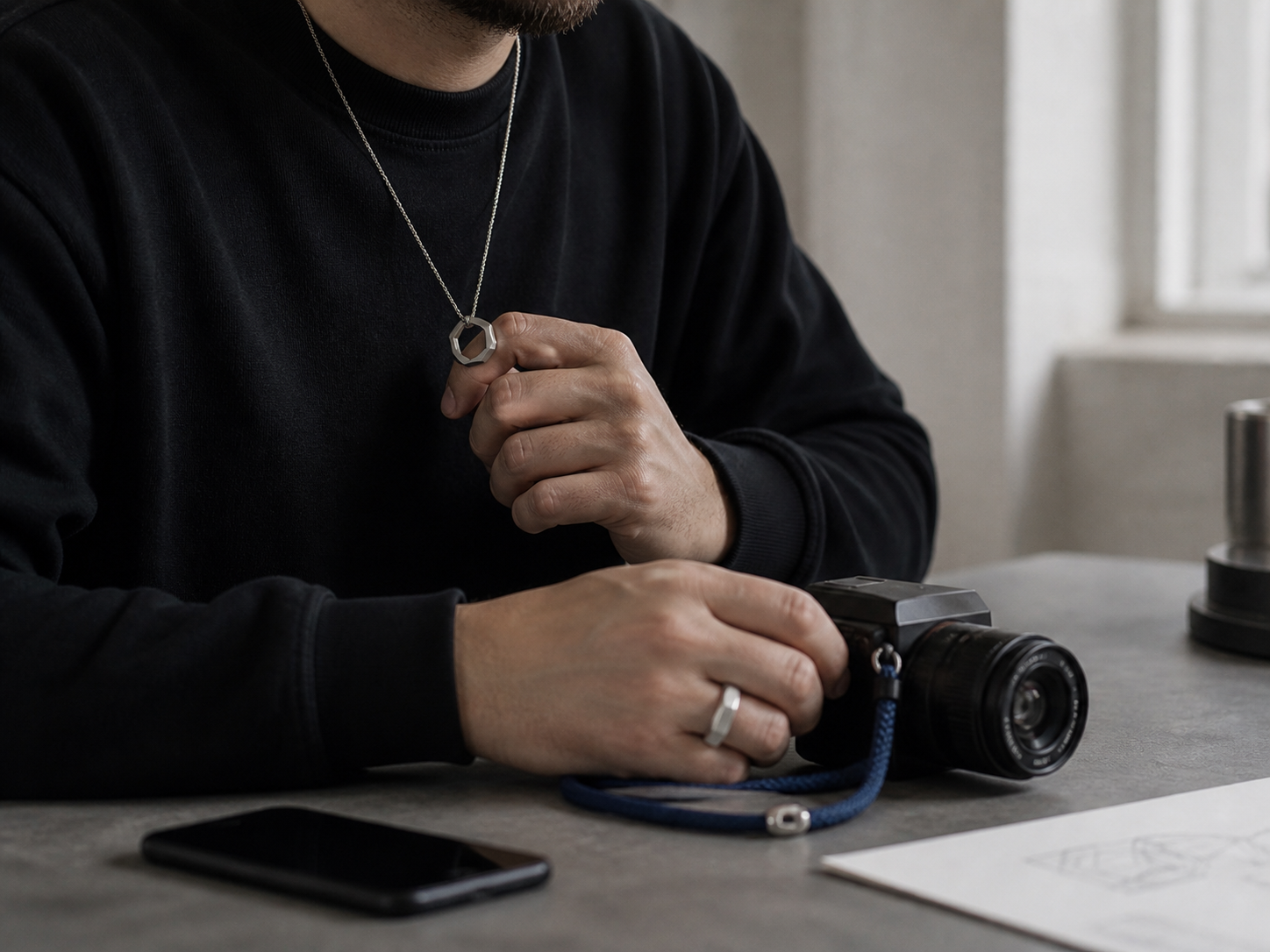 Person mit silbernem Ring und Kette hält eine Kamera neben einer Schmuckskizze